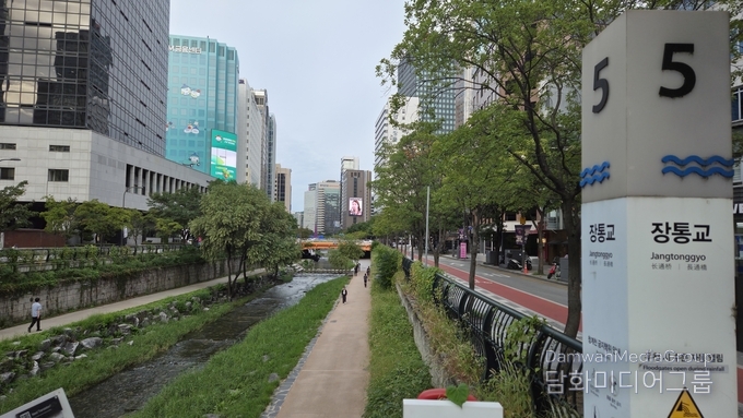 사진은 서울 도심 속 힐링 공간인 청계천 장통교 부근 전경을 담고 있다. 화면 오른쪽에는 ‘장통교(Jangtonggyo)’ 안내 기둥(5번 표식)이 자리해 위치를 알려주고 있으며, 5번 출구를 나오면 곧장 보신각 방향 도심 거리가 이어진다. 청계천 물길은 왼쪽으로 흐르고, 산책로를 따라 시민들이 여유롭게 걷는 모습이 포착되었다. 양옆으로는 고층 빌딩 숲이 들어서 있어 전통과 현대가 공존하는 서울의 풍경을 잘 보여준다. 특히 이 지점은 보신각에서 불과 60미터 거리에 있어 접근성이 뛰어나며, 청계천 산책과 종각 일대의 도심 문화를 함께 즐길 수 있는 최적의 장소임을 보여주고 있다.